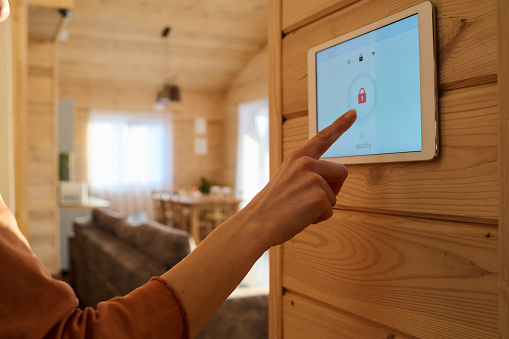 Hand of young woman or housewife pointing at screen of alarm system panel with lock sign hanging on wooden wall in living room