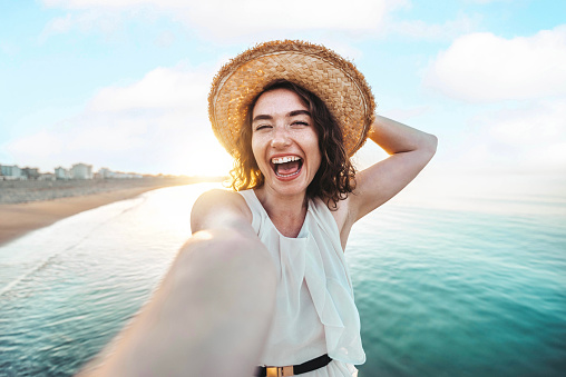 Happy beautiful young woman smiling at the beach side - Delightful girl taking selfie picture with smart mobile phone device outside - Healthy lifestyle concept with female enjoying sunny day