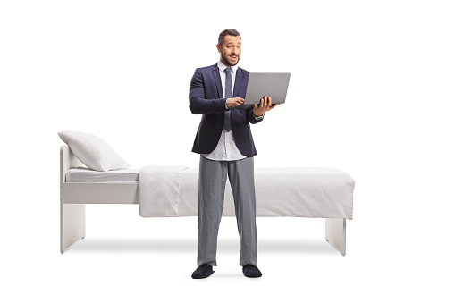 Man wearing a suit and bottom pajamas and holding a laptop computer in front of a bed isolated on white background