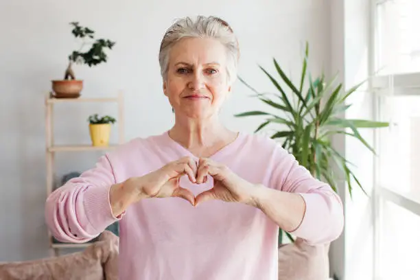 Middle-aged mature woman in living room connected fingers showing heart symbol close up, people cardiovascular disease prevention treatment, health check-up, cardio vitamins, sign of kindness and charity