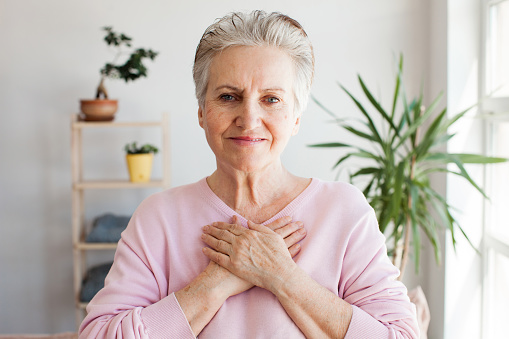 Portrait of happy adult elderly female volunteer holding folded hands on chest, looking at camera. Kind smiling middle aged woman feeling thankful, showing appreciation, gratitude believe charity concept
