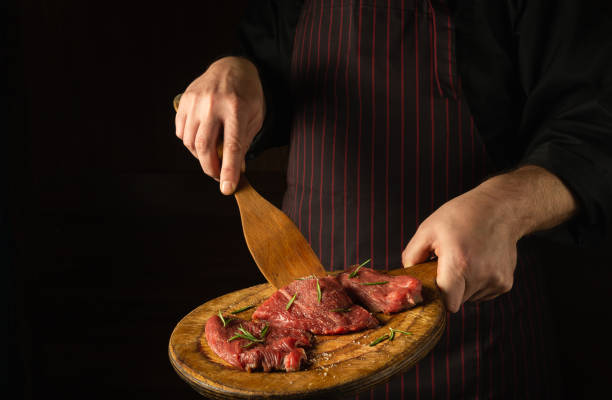 istock The chef turns over raw beef steaks with a wooden spatula before barbecue. Preparation for roasting meat with rosemary and spices. Black space for hotel recipe or menu. 1492571302