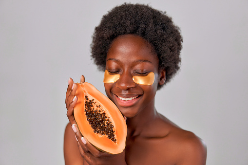 African American young woman with afro hair and perfect smooth healthy skin using eye patches and holding papaya in hands isolated on gray background. Skin care and spa treatments.