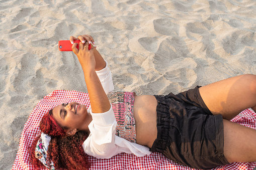 Latin woman checking her cell phone while lying on a blanket on the beach