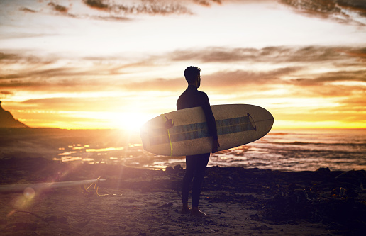 Back, surf and sunset view with a man on the beach during summer for travel, vacation or recreation. Sky, nature and surfing with a male surfer on the coast for a water sports hobby while on holiday