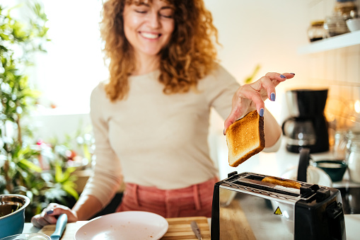 Smiling young woman making breakfast toast bread with toaster at home kitchen