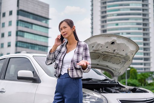 Young woman standing next to her car calling for assistance after car breakdown at roadside of urban city