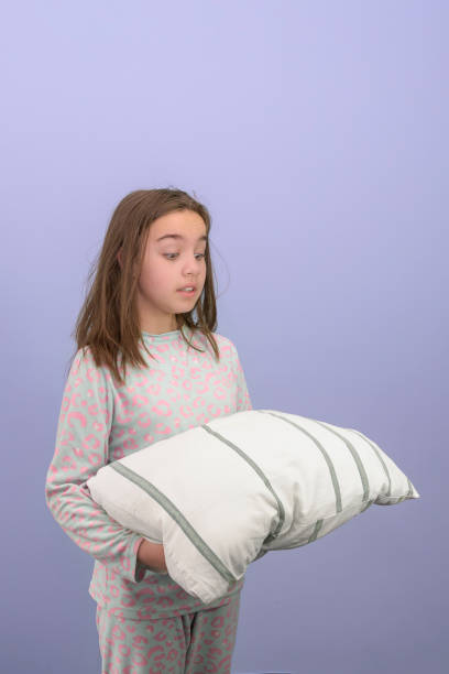 パジャマを着た10代の少女がベッドの準備をしています。少女は枕を切望して見ています。スタジオ写真、垂直、薄紫色の背景。 - longingly ストックフォトと画像
