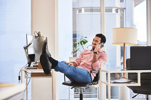 Relax business man with his feet up on desk on a phone call with job confidence and successful career. Asian CEO, boss or professional person relaxing in office and talking on smartphone