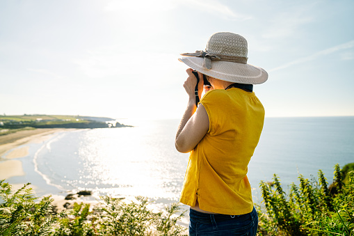 A casual blonde tourist, donning a bright yellow tee and a colorful hat, captures a scenic coastal landscape from a cliff adorned with lush greenery in the midday light.