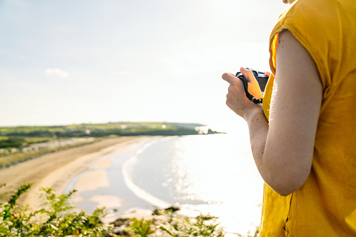 Unrecognizable tourist in casual yellow tee capturing coastal landscape with camera on lush green cliffside in midday light.