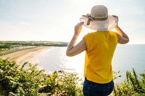 Unrecognizable casual-dressed tourist girl, wearing a colorful yellow tee, touching her hat, gazing at coastal scenery from green-clad cliff with noonday sunlight.