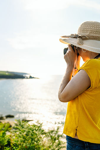 Casual close-up of a vibrant blonde tourist in a colorful yellow tee and hat, capturing coastal scenery from a lush green cliff in midday light.