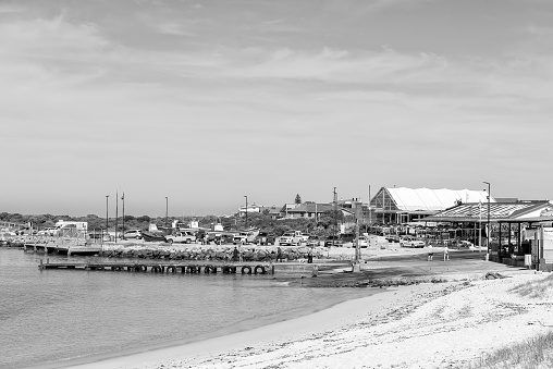 Struisbaai, South Africa - Sep 21, 2022: View of the harbor in Struisbaai, in the Western Cape Province. People, boats, vehicles and the waterfront are visible. Monochrome