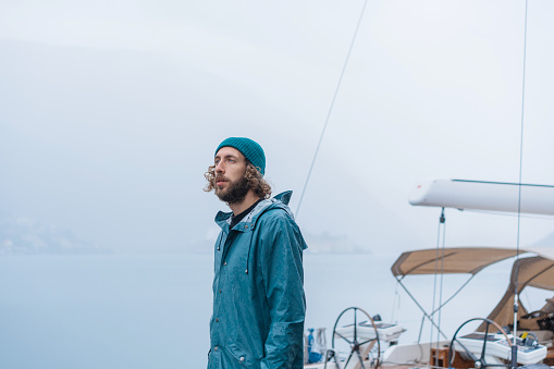 Man in durable blue raincoat standing  near the sailing  boat during the rain