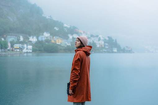 Woman in orange  raincoat standing on the background of Odda town and fjord during the rain