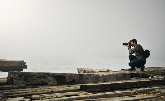 Travel photographer, man at sea and taking photo with digital camera on the rocks. Outdoor photography, ocean view or beach environment and male on knee recording the ocean view on a high peek