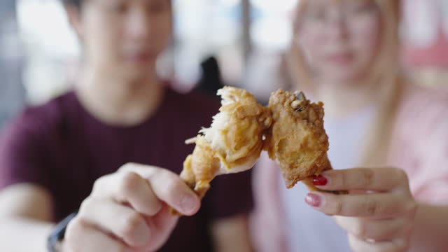 Defocused shot of young Thai couple holding fried chicken giving toast to each other and taking a big bite