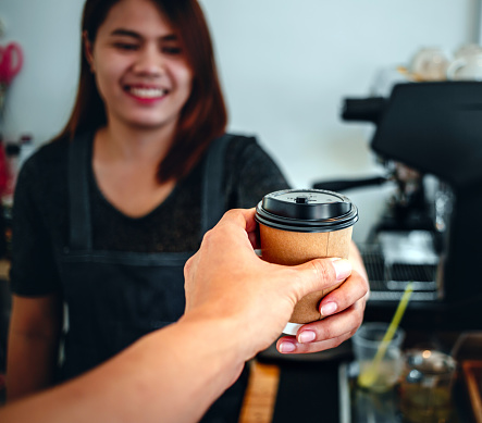 A friendly smiling female barista is holding in hands hot coffee in a takeaway paper cup submitted to the customer in a coffe shop. Small business concept
