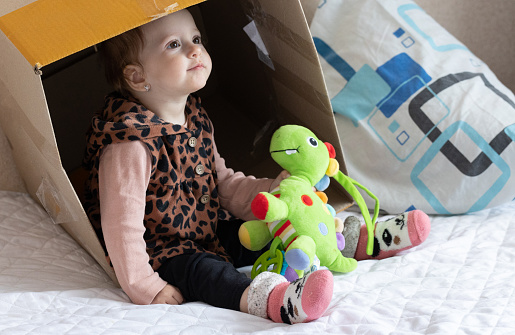 Little girl plays inside a cardboard box on the bed in the bedroom.