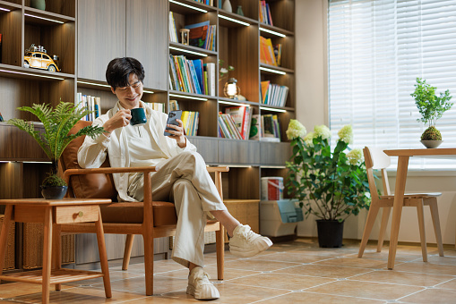 An Asian young man wearing white casual clothing. Sitting in the living room at home to rest. Hold your phone and watch.