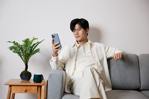 An Asian young man, dressed in white casual clothing, sat on the sofa in the living room, browsing media on his phone. He is very relaxed.