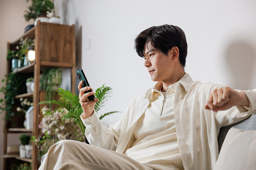 An Asian young man, dressed in white casual clothing, sat on the sofa in the living room, browsing media on his phone. He is very relaxed.