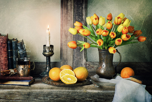 Classic still life with bouquet of tulips flowers placed with fresh oranges, scarf, vintage books and illuminated candle on rustic wooden table.