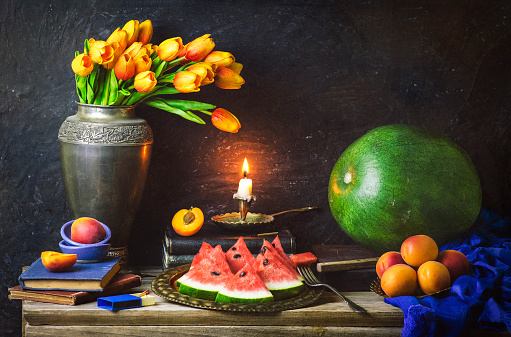 Classic still life beautiful bouquet of tulips flowers placed with old books, illuminated candle, blue scarf,  fresh watermelon and apricots on rustic wooden background.