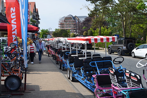 De Haan, West-Flanders, Belgium- May 21, 2023: walking female senior friends walking on footpath. Family pedal go-carts striped marquee roof stationary in a row next to sidewalk to rent on a sunny day