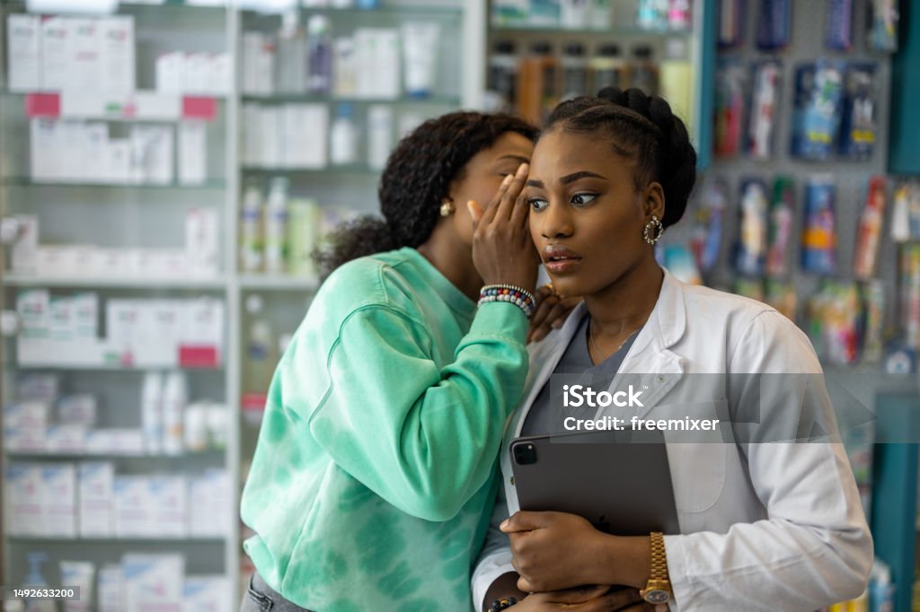 Woman whispering to a pharmacist Black female pharmacist listening what a female customer is whispering in her ear in pharmacy. Whispering Stock Photo