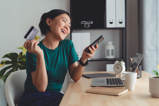 Asian woman happy with online shopping, easy online payment, holding credit card and using smartphone for shopping online at home office.