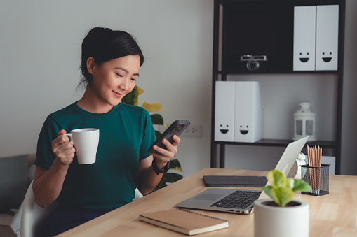 Asian woman chill with relax time take a break drinking warm coffee while using smartphone sitting at home office.