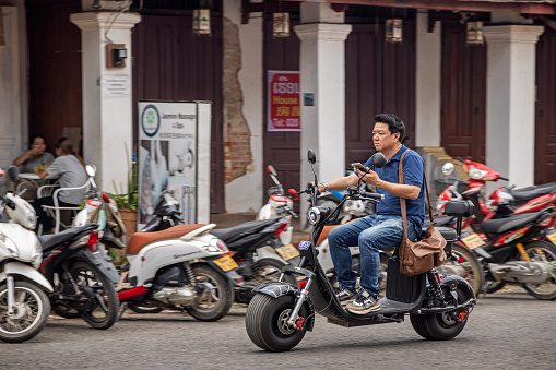 Luang Prabang, Laos - March 18th 2023: One man with a mobile phone on a electric motorbike