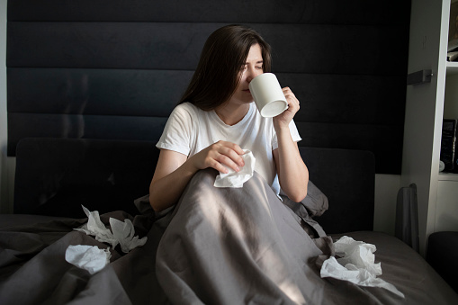 Woman lying in bed using a handkerchief. Lots of scattered handkerchiefs. Woman drinks tea from a white mug hot tea or water. Concept: illness, cold, coronovirus, flu epidemic