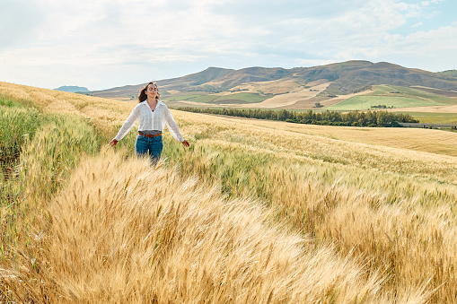 Woman walking in golden wheat field in hot summer sun and blue sky with white clouds with mountains hill landscape in background.