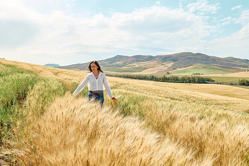 Woman walking in golden wheat field in hot summer sun and blue sky with white clouds with mountains hill landscape in background.