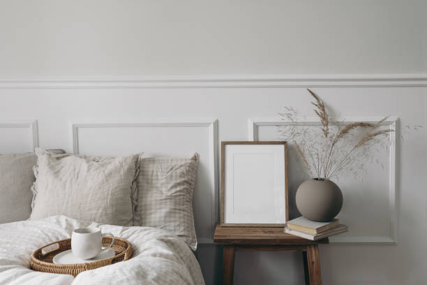 istock Elegant boho bedroom interior. Vertical wooden picture frame mockup on old bedside table. Cup of coffee. Ball vase with dry grass. Beige linen throw bedding. White wall background. Scandinavian home. 1492643059