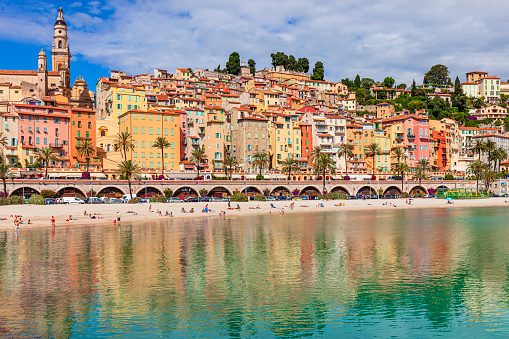 People enjoying a sunny spring day on the Plage des Sablettes of Menton, a famous seaside resort on the French Riviera, with the colorful buildings of the old town reflected in the water.