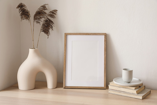 Blank wooden vertical picture frame mockup. Vase with dry reed, grass on table, desk. Cup of coffee, books. Elegant interior, home office. Decorative boho still life photo, artistic poster display.