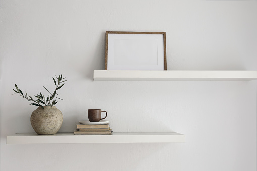 Modern interior still life. Two floating shelves. Blank wooden picture frame mockup template. Vase with olive tree branches, old books, cup of coffee. Modern Mediterranean home, white wall background.