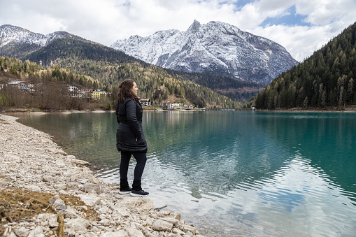 Woman in black clothes standing on lakeside of Auronzo Lake, in Auronzo di Cadore village; Snow peaked mountains on background; Belluno, Dolomites, Italy