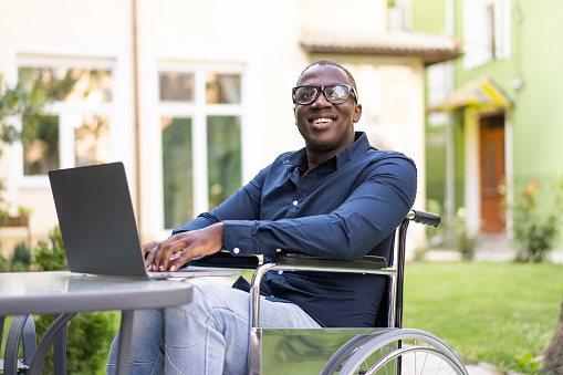 An Anglo-American man is using his laptop, sitting in a wheelchair while he recovers from an injury, working outside in a garden on a sunny day