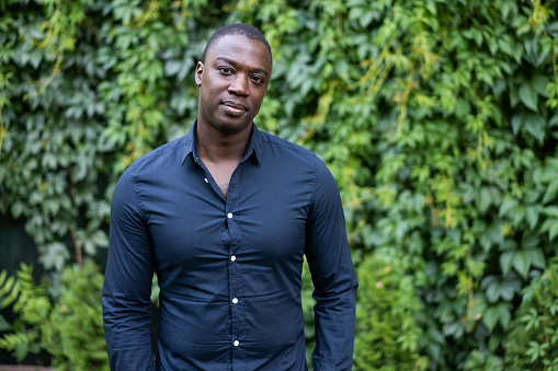 Portrait of an African-American man from the waist up, wearing a smart shirt and looking seriously at the camera, with an ivy-covered wall in the background