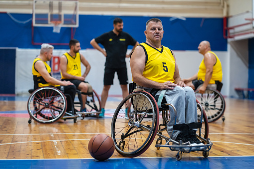 An older athlete with disabilities sitting in a wheelchair wearing a basketball uniform looking at the camera with basketball on the floor by his wheels, team players in wheelchairs in the background listening to their coach
