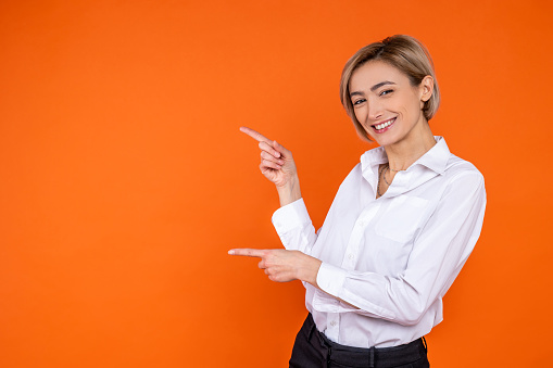 Delighted woman wearing white official style shirt pointing at advertisement area isolated over orange background.