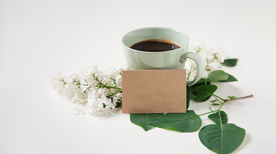 Black coffee in pastel green cup with white lilac flowers and brown empty note on white background.