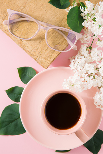 High angle view of black coffee in pastel pink cup, papyrus notebook, pastel pink reading glasses, decorated with green leaves and white lilac flowers on pink background.