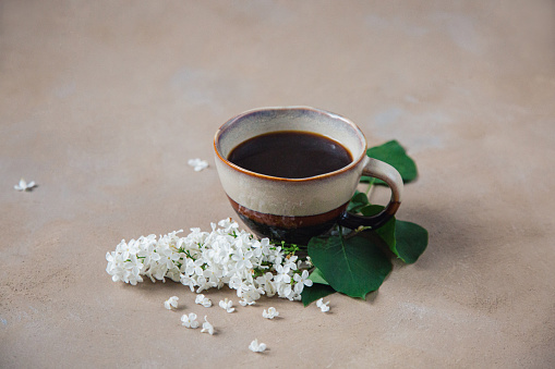 Black coffee in beige-brown cup decorated with green leaves and white lilac flowers on rustic brown-beige stone texture table.