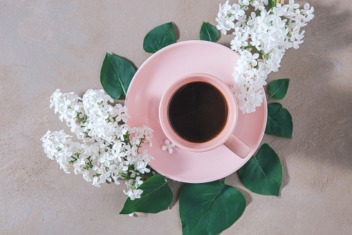High angle view of black coffee in pink cup surrounded with green leaves and white lilac flowers on pink background. Vintage processing.